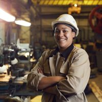 Waist up portrait of mixed-race female worker posing confidently while standing with arms crossed in factory workshop